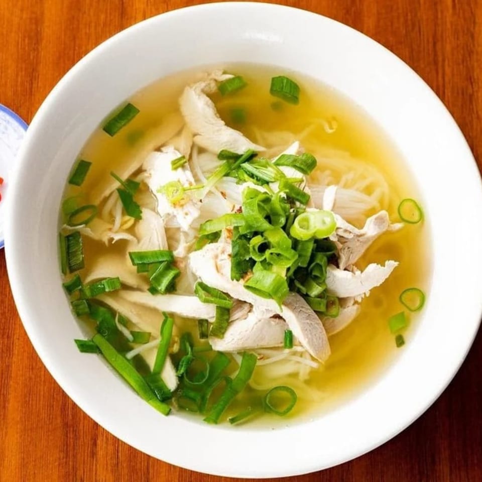 A white bowl of Pho-Chau chicken pho with broth, noodles, and green onions.