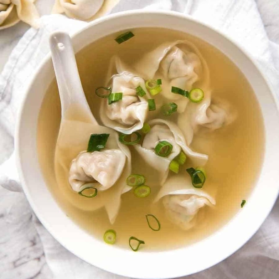 Overhead view of a white bowl of Pho-Chau wonton soup with green onions and a spoon.